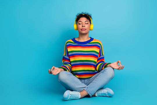 Young woman meditating in a bright rainbow striped sweater with yellow headphones against a blue studio background