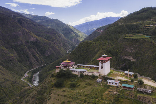 Aerial view of a fortress Dobji Dzong perched atop a hill, overlooking a river winding through lush, green valleys under a partly cloudy sky, Raga, Paro, Bhutan.