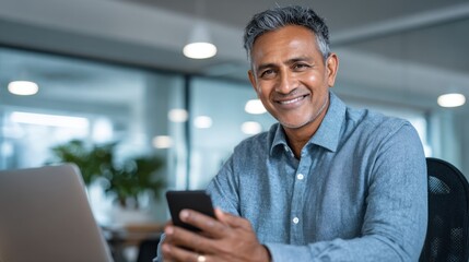 Smiling Indian businessman in blue shirt holding smartphone with laptop and plant in office