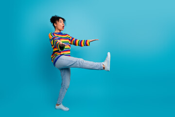 Young woman in a rainbow striped sweater kicks playfully against a blue backdrop conveying energy...