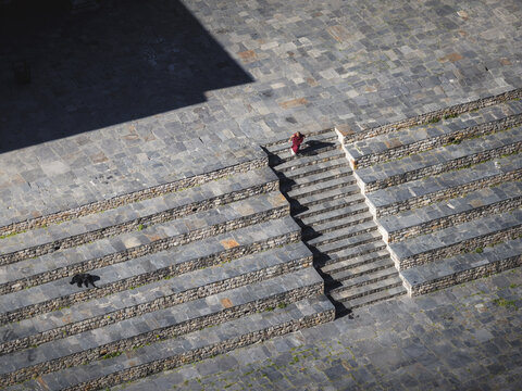 Haa, Bhutan - 26 September 2025: Aerial view of a lone monk in vibrant red robes ascending the stone steps within the ancient Haa Dzong, where shadows dance on the textured courtyard.
