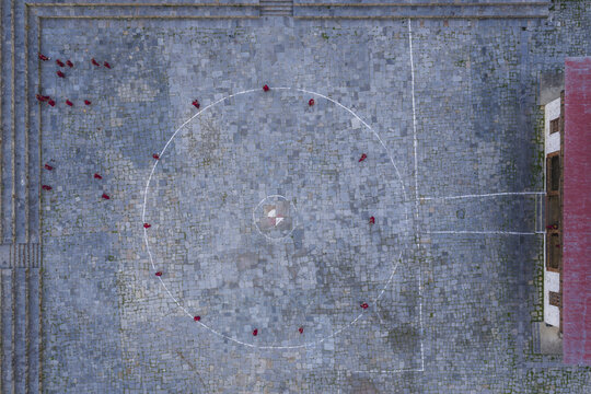 Aerial view of monks in vibrant red robes circle a central point within the Haa Dzong's textured stone courtyard, Haa, Bhutan.