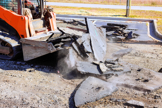 Construction skid steer machinery is breaking up asphalt in parking lot while dust rises into air