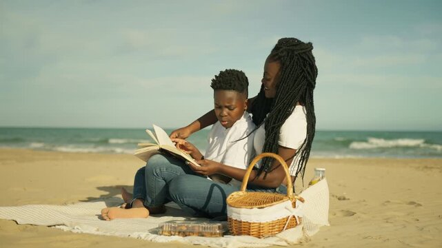 Loving african american mother and young son enjoy sunny day at beach, read book together while  picnic on  sand with beautiful ocean waves in background