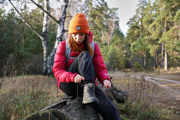 A young female hiker is lacing her trekking boots while sitting on a log in the forest. Hiking in the forest in autumn