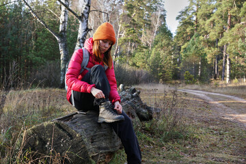 A young female hiker is lacing her trekking boots while sitting on a log in the forest. Hiking in the forest in autumn