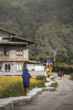 View of a person walking along a winding path lined with prayer flags and yellow fields near a multi-story building, Sopsokha, Punakha, Bhutan.