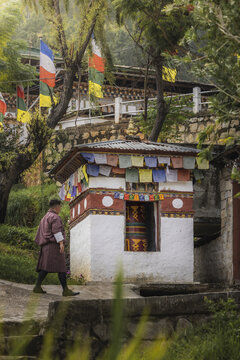 View of a man in traditional Bhutanese clothing walks past a prayer structure adorned with colorful flags in a tranquil setting, Sopsokha, Punakha, Bhutan.
