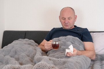 Man sitting in bed under a blanket, holding a tissue and appearing to have cold or flu symptoms