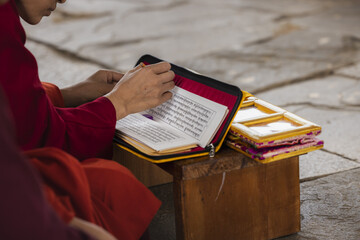 View of a monk, bathed in warm hues, engrossed in ancient scripts within a vivid yellow case, seated serenely on a wooden stool, Wolakha Nunnery, Punakha, Bhutan.