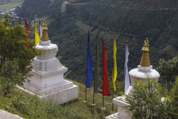 View of white stupas and vibrant prayer flags fluttering against a backdrop of lush green mountains, offering a serene contrast, Wolakha Nunnery, Punakha, Bhutan.