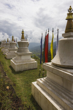 View of white stupas with golden spires stand elegantly against the vivid prayer flags fluttering in the wind, overlooking the lush valley, Wolakha Nunnery, Punakha, Bhutan.