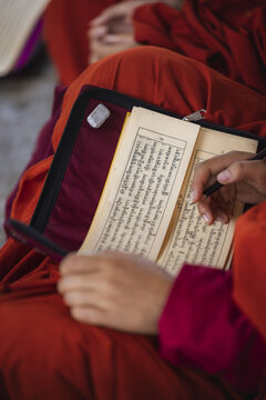 View of a monastic's hands gently holding an open book with complex scripts, poised with a pen, contrasting against the deep red robes, Wolakha Nunnery, Punakha, Bhutan.