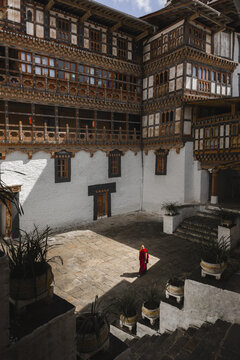 Trongsa, Bhutan - 22 September 2025: View of a lone monk in vibrant red robes walks across the sun-drenched courtyard of the Trongsa Dzong, its intricate wooden balconies casting shadows.