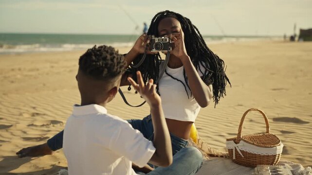 Young woman with dreadlocks use vintage camera to photograph smiling son on sunny sandy beach during relaxed family picnic and seaside vacation moments