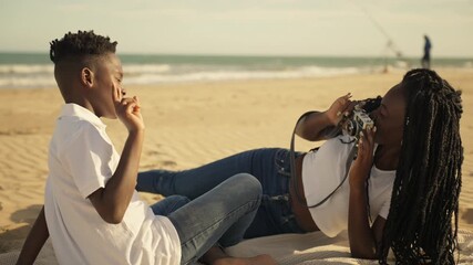 Happy african american woman with dreadlocks lying on blanket at beach, taking photos of young boy with vintage film camera as he poses playfully for during sunny day by sea
