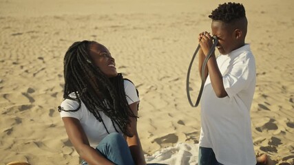 Happy young african american son taking pictures of his smiling mother with vintage camera while having fun picnic together on sunny sandy beach during their family summer vacation