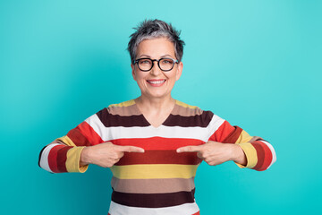 Aged mature woman with short gray hair and glasses smiles and points to her colorful striped sweater turquoise background for lifestyle advertising use