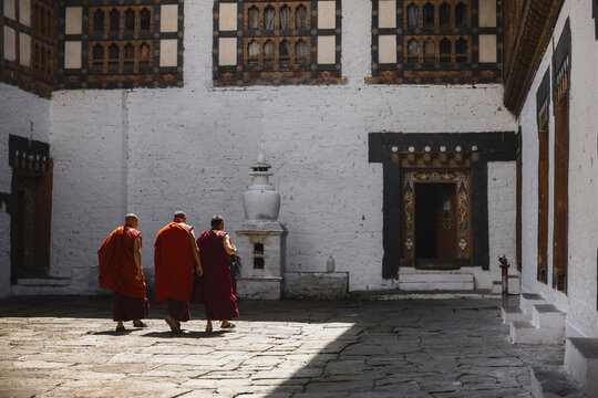 Trongsa, Bhutan - 22 September 2025: View of crimson-robed monks traverse the sun-drenched courtyard of Trongsa Dzong, its white walls juxtaposed against ornate dark wooden window frames.