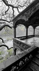An ornate, weathered stone balcony with intricate carvings and columns overlooks a calm, misty lake. Bare, gnarled tree branches frame the scene, suggesting a c