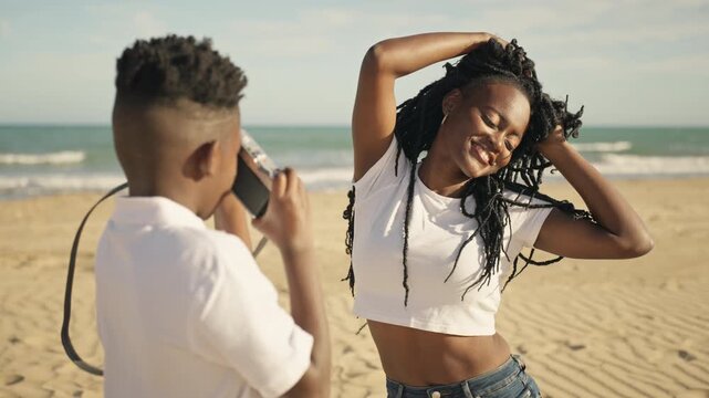 Cheerful black woman with long dreadlocks posing for little brother, who take photos of her with vintage camera during their happy summer vacation at sunny seaside