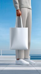 A person stands outdoors, holding a large white tote bag. They are wearing light-colored clothing and white sneakers. The background features a bright blue sky