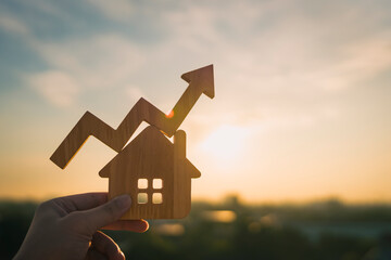 Hand holding wooden house with rising arrow graph at sunset, symbolizing real estate investment growth, property market trends, mortgage rates and housing price increase in financial concept.