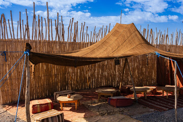 Traditional outdoor tent with woven walls, wooden tables, and vibrant cushions under a blue sky. Agafay Desert, Marrakesh-Safi, Morocco