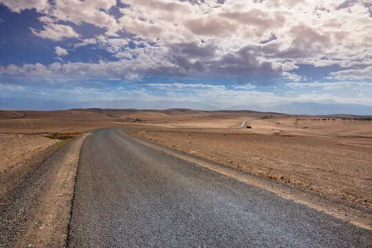 Empty winding road through vast desert landscape under a blue sky with scattered clouds. Agafay Desert, Marrakesh-Safi, Morocco