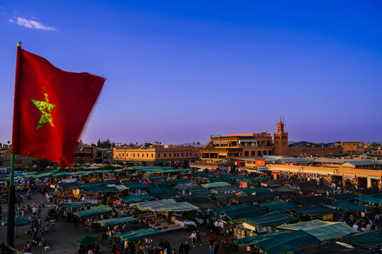 Vibrant city market at dusk with a red flag and historical architecture. Marrakesh, Morocco