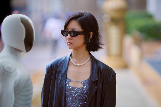 Woman in smart sunglasses with short hair and pearls facing a humanoid AI robot outdoors. Shanghai, China