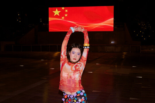 Woman poses in vibrant outfit at night with illuminated Chinese flag billboard behind her. Shanghai, China