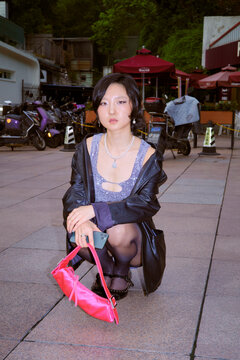 A young adult crouching on a paved area, holding a pink purse, with bikes in the background. Shanghai, China