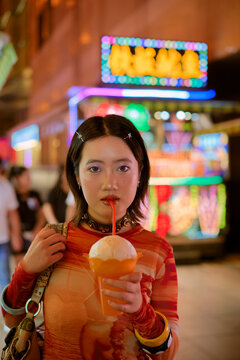 Young woman in a vibrant outfit drinks from a cup with a colorful city backdrop. Shanghai, China