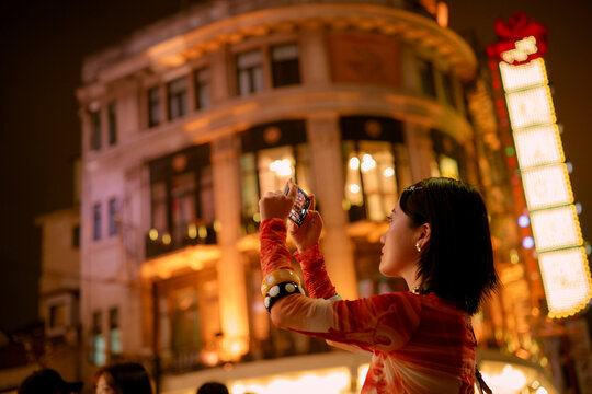 Woman in vibrant clothes photographs illuminated building at night in bustling city street. Shanghai, China