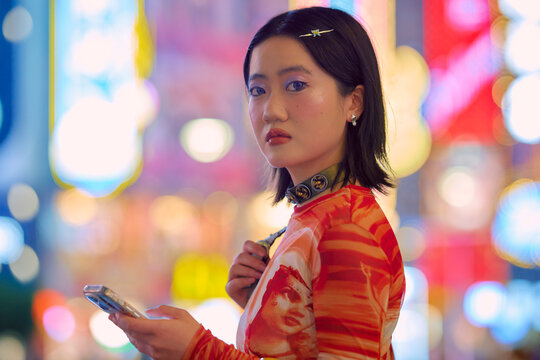 Young woman with phone stands against a vibrant city nightlife backdrop. Shanghai, China