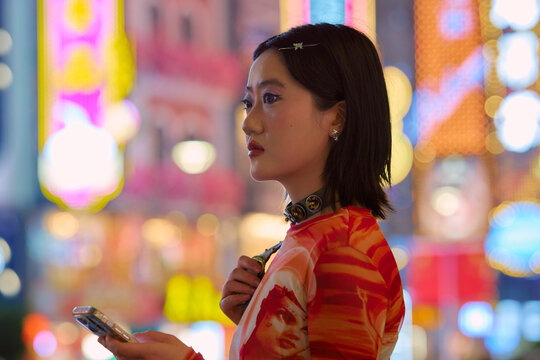 Woman with phone in hand stands against vibrant, colorful city nightlife background. Shanghai, China