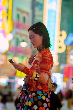 Young woman in vibrant clothing checks her phone amid colorful nightlife. Shanghai, China