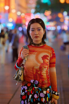 Young woman in a colorful outfit stands on a vibrant, busy city street at night. Shanghai, China