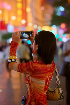 Woman capturing colorful city lights at night on a smartphone, showing a bustling urban scene. Shanghai, China