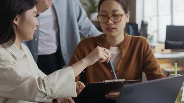Handheld shot of Asian coworkers talking about details of project holding folder with document and using laptop at desk in modern office