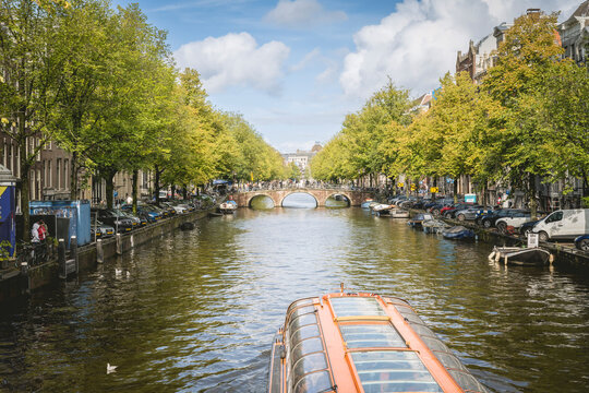 Canal in Amsterdam lined with boats and trees, featuring a bridge under a partly cloudy sky. Amsterdam,. Netherlands