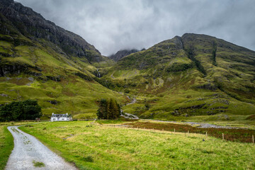 Serene landscape of a green valley with a white cottage and majestic mountains under a cloudy sky. Glencoe, Scotland