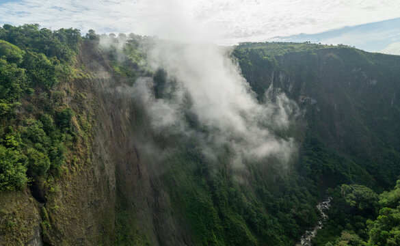 Misty cliffs covered in lush green vegetation overlook a deep valley under a partly cloudy sky. San Agustin, Colombia