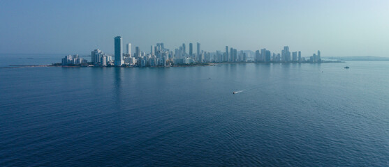 Skyline of a city by the sea with a boat traversing the calm blue water under a clear sky. Cartagena, Colombia