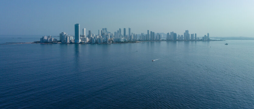 Fototapeta Skyline of a city by the sea with a boat traversing the calm blue water under a clear sky. Cartagena, Colombia