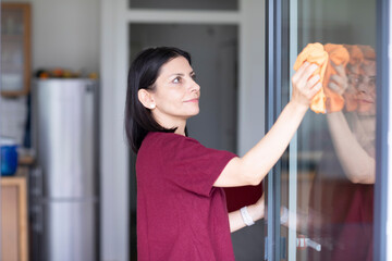 Woman in red shirt cleaning a glass door with an orange cloth in a modern kitchen. Germany