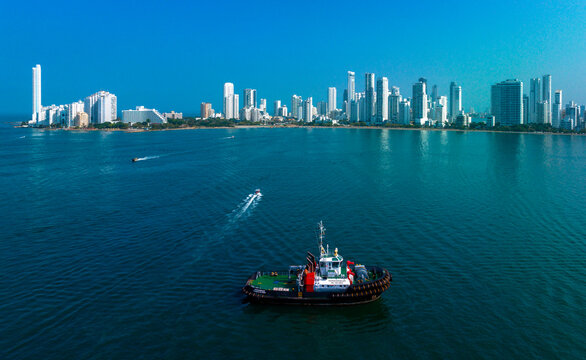 A tugboat moves across a calm blue sea with a city skyline in the background under a clear sky. Cartagena, Colombia