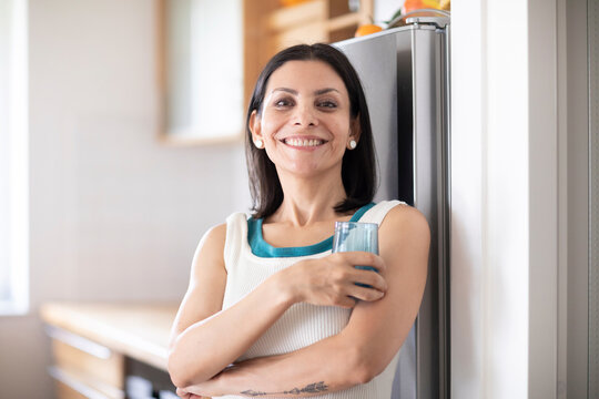 Smiling woman with long dark hair holding a glass of water in a bright kitchen. Germany