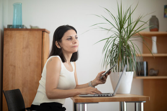 A woman in a white top sits at a desk using a laptop and holding a phone. Germany
